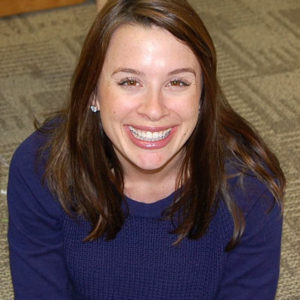 A young woman with a dark sweater and long brown hair smiles and looks up at the camera